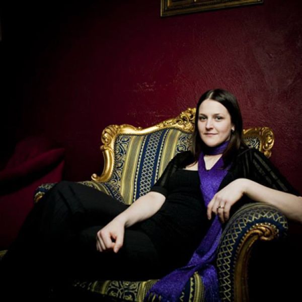 A person wearing black clothing and a purple scarf reclines on an ornate blue-and-gold armchair against a deep burgundy wall.
