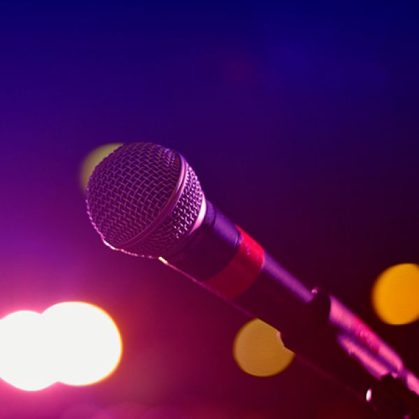 A close-up of a microphone on a stand is illuminated by pink and purple stage lighting, with blurred concert lights creating a colourful background.