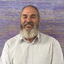 Head-and-shoulders portrait of proud Bidjara Gunggari Mhurdi man, Lindsay Hardy, with a grey beard wearing a light-coloured collared shirt. Lindsay is standing in front of a textured blue and purple background, with the portrait cropped from the chest up.