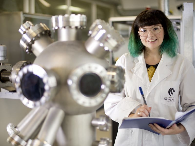 Female student with blue hair standing in physics lab smiling at camera, holding a notepad. 