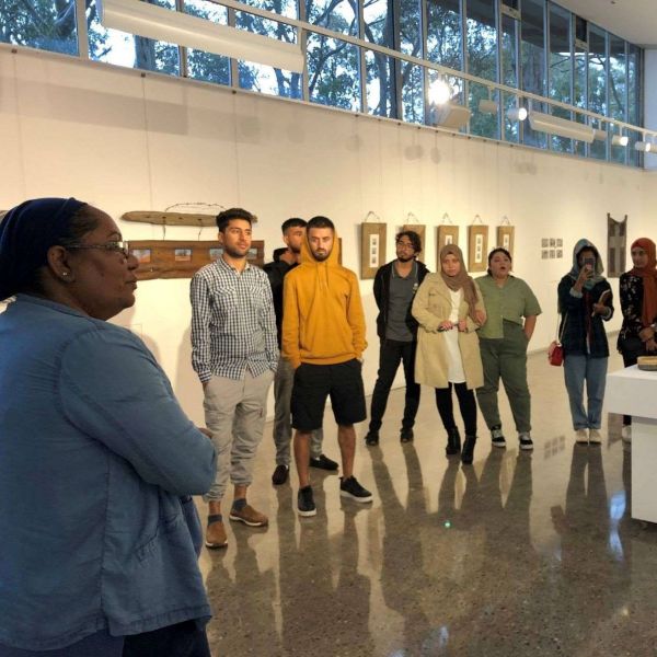 A woman speaking in an art gallery to a group of young men and women. Young refugee adults learn about Aboriginal resilience and survival 