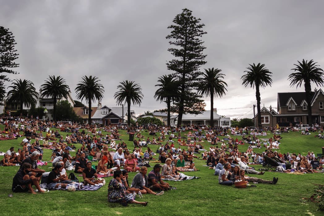 A large group of people siting in a park picnicking watching a performance. the performance can not be seen in this picture.