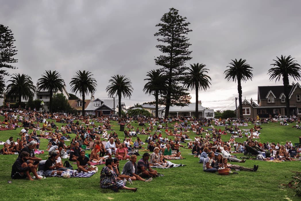 A large group of people siting in a park picnicking watching a performance. the performance can not be seen in this picture. 