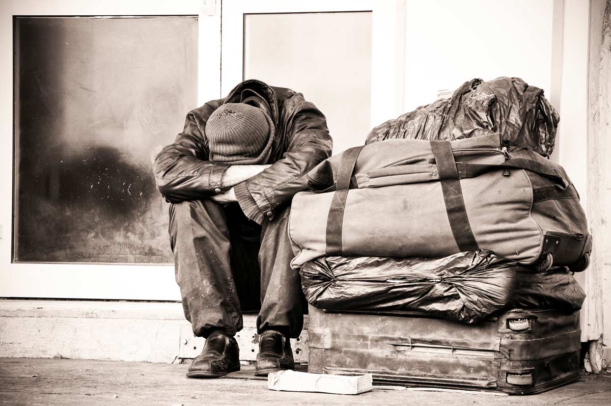 Sepia toned image of a homeless person sitting on a street with a pile of luggage.