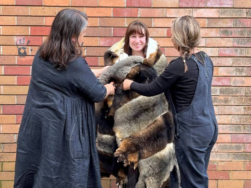 Two female adults wrapping a high school student in an ankle-length possum skin cloak