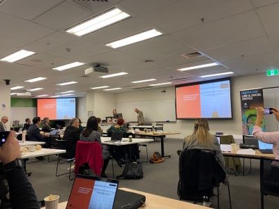Terry Carney presenting at the Algorithmic Justice Symposium. The audience is pictured sitting at desks facing towards Terry Carney.
