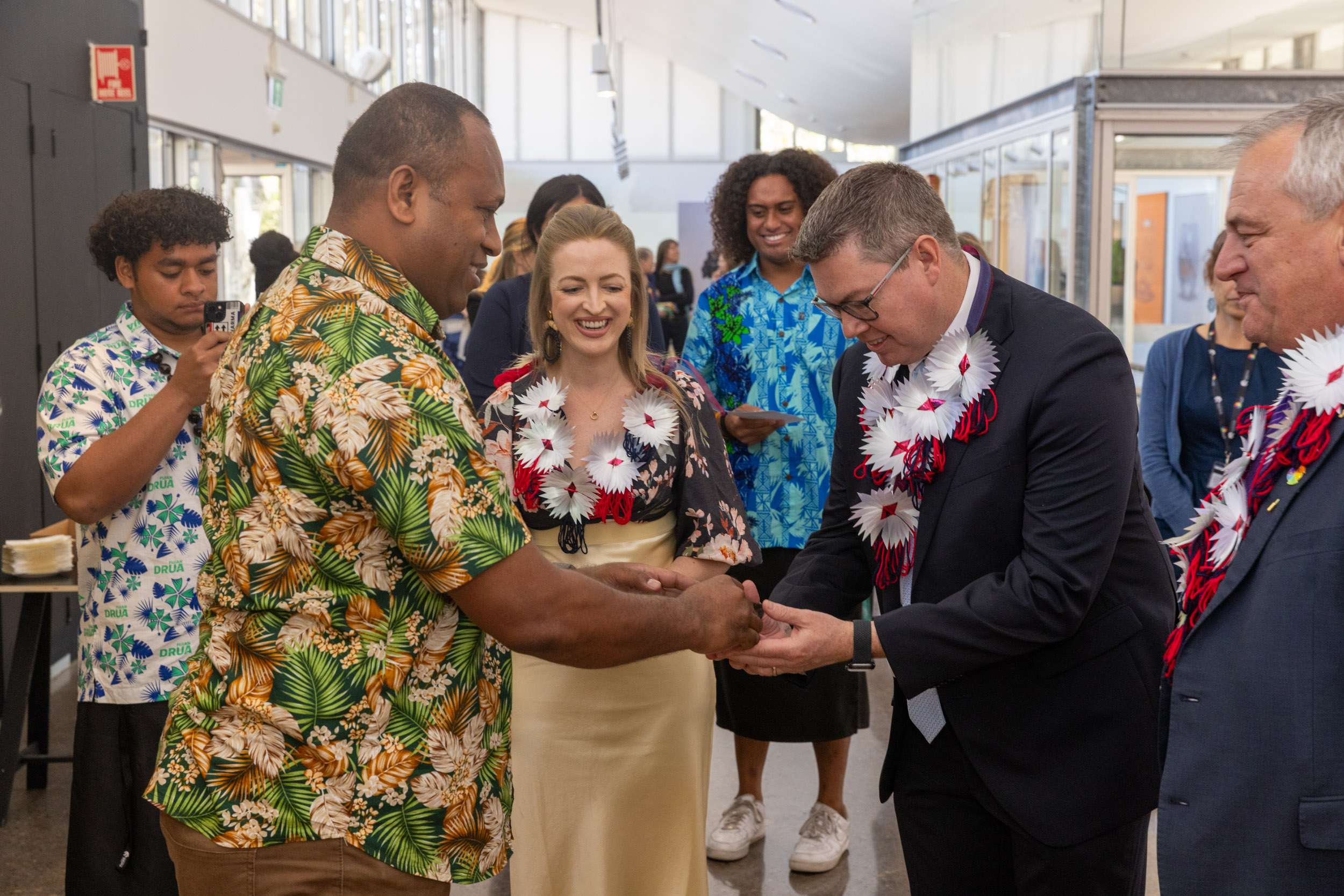 A man wearing a green floral Pacific shirt hands another man wearing a suit a flower lei. A woman with a lei stands in the middle and two men can be seen in the background taking photos