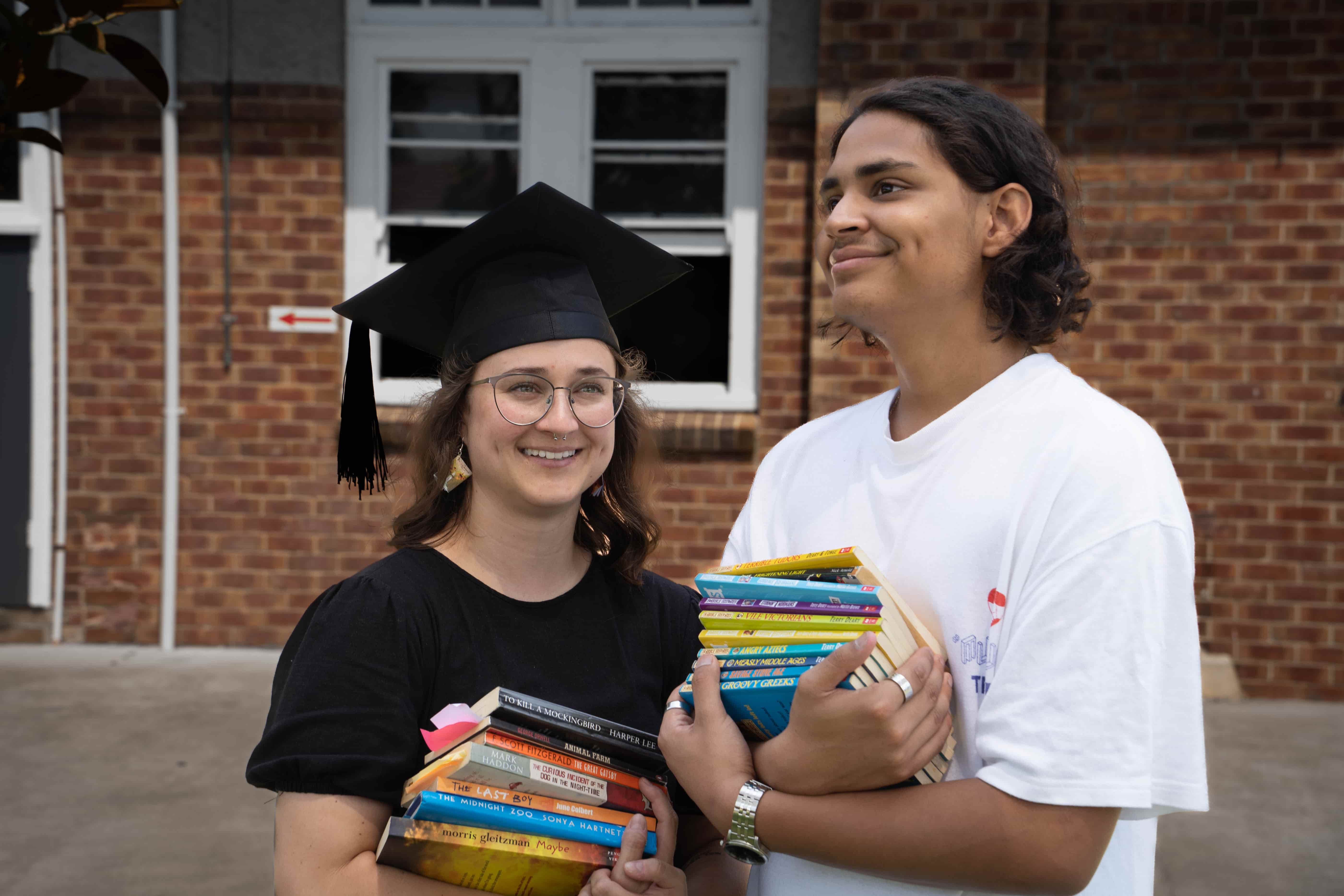 Kat and Tom Kat pictured in her graduation cap holding a stack of books with student, Tom looking excited and hopeful