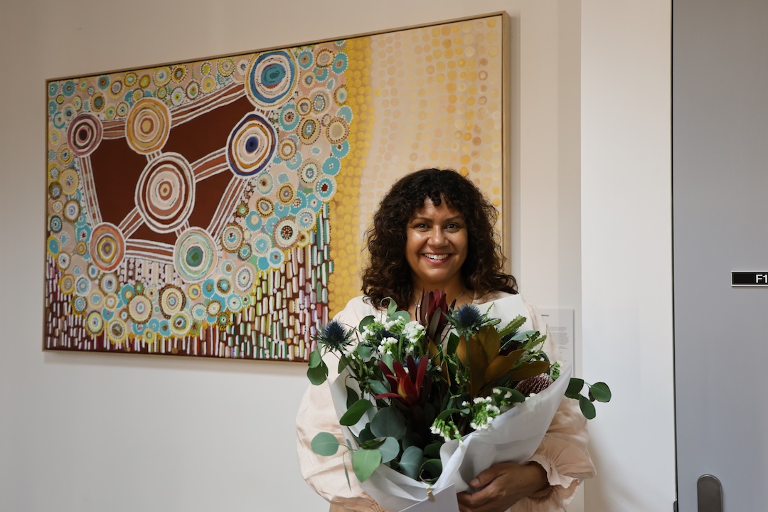 Artist Josie Rose poses with her artwork Wild Flowers at the official unveiling in Port Macquarie. She is holding a bunch of native flowers and poses smiling in front of the piece.