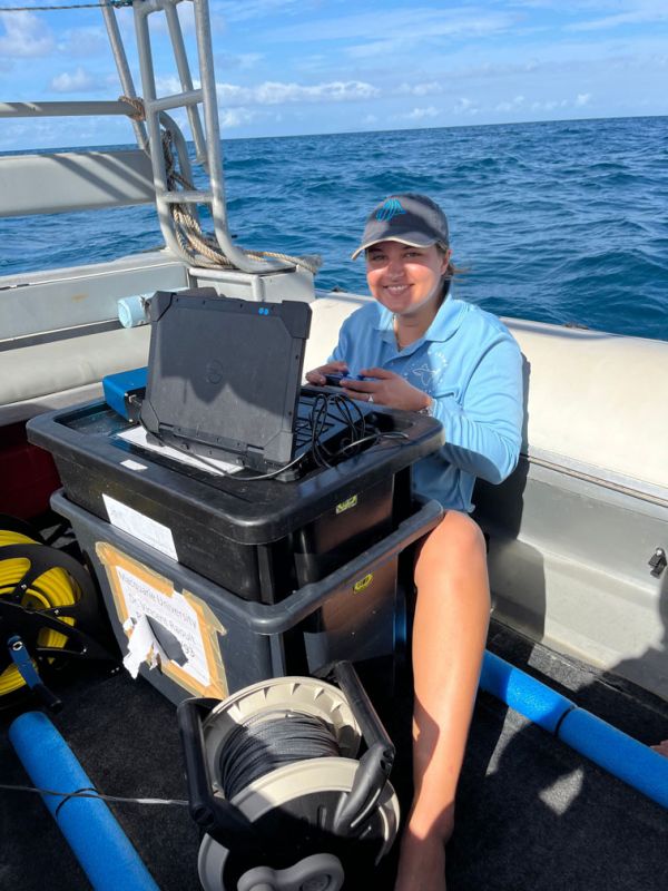 Kristen operating a laptop connected to marine equipment on a boat under a clear sky.