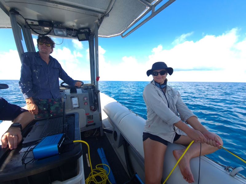 Two people on a boat under a clear blue sky, one steering and the is Kristen seated on the side, both wearing sunglasses and hats, with open ocean in the background.