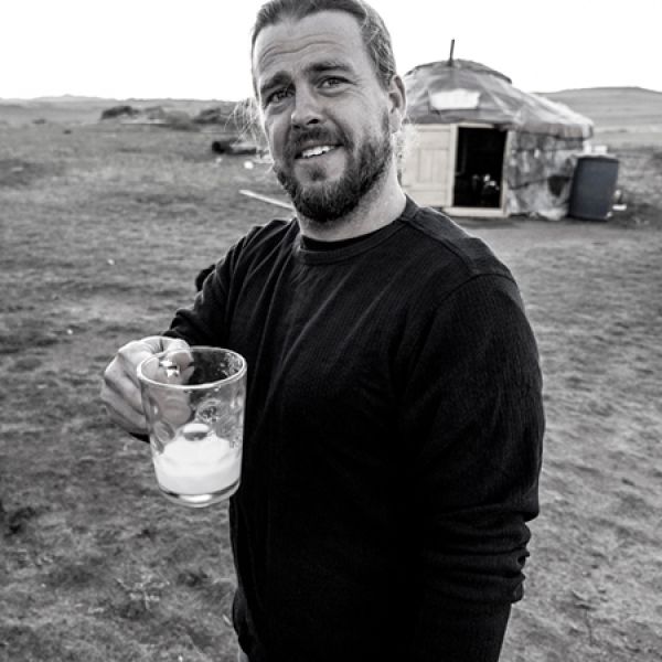 A black-and-white photograph of a person standing outdoors on open land, holding a glass with a light-colored drink, with a small round tent-like structure in the background.