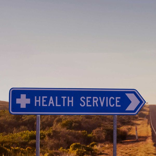 A road sign with text 'Health Service' along a highway road with desert terrain.. Research to Improve Health in Regional Communities.