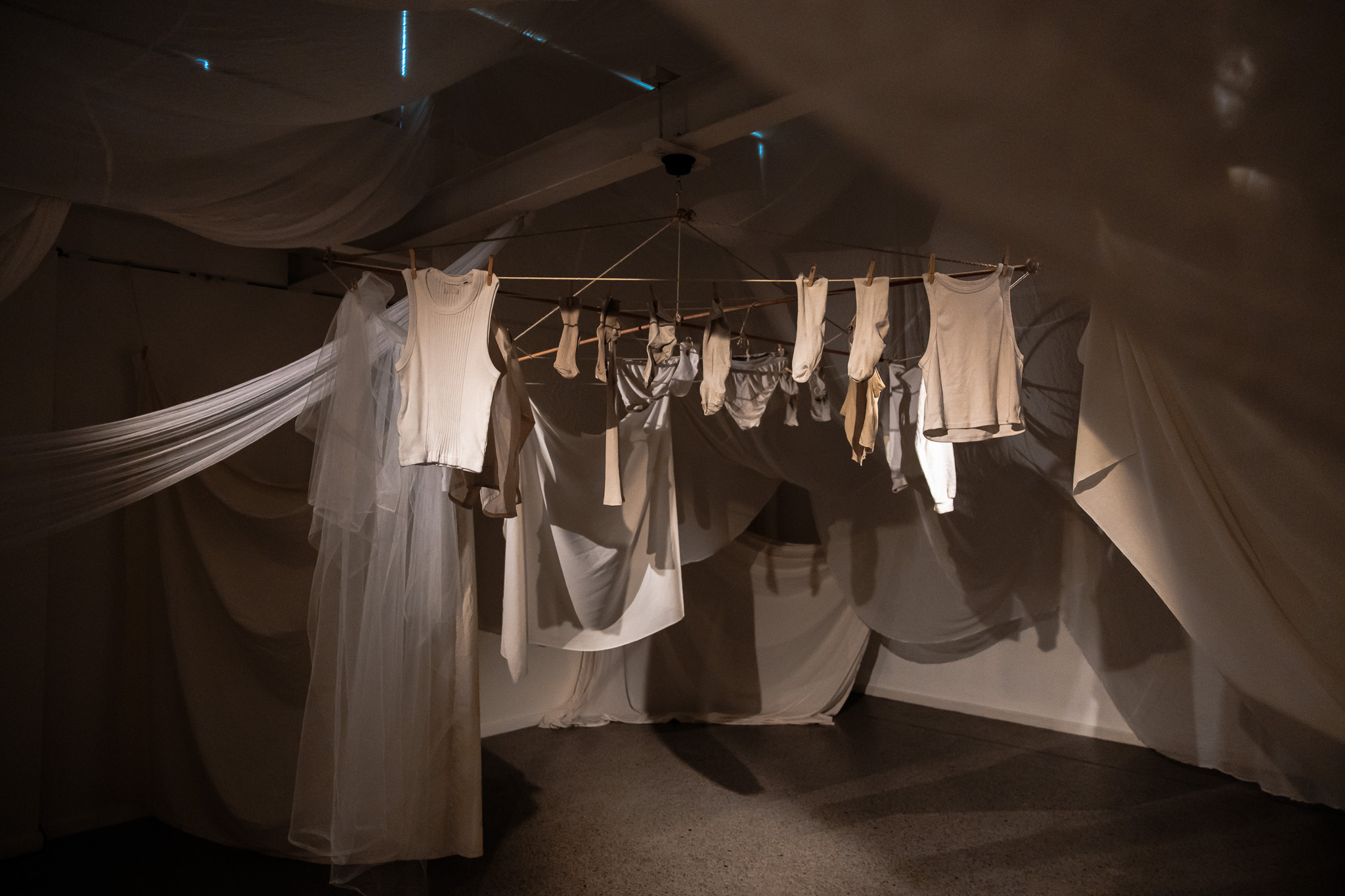 A n image of a darkened gallery space with pale-coloured washing on a line