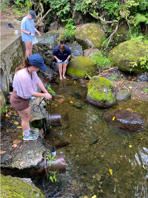 Three people stand on rocks in a small body of water, holding equipment.