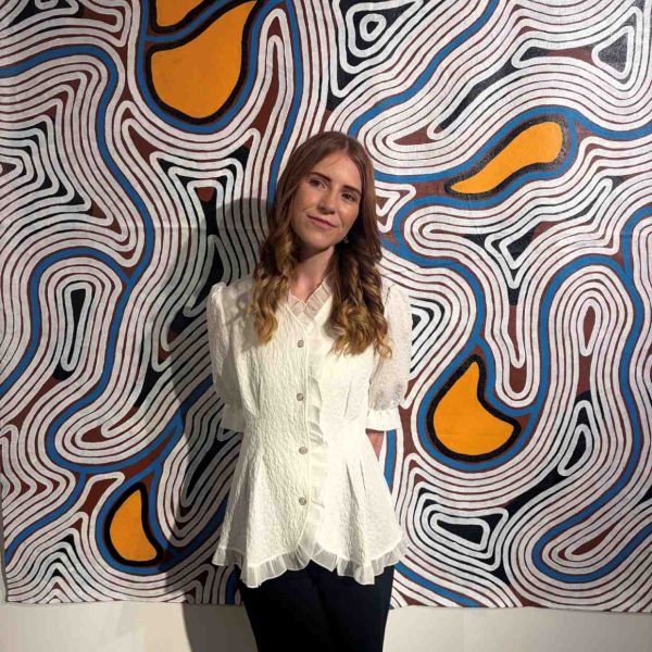 Steph smiling at camera with hands behind her back and standing in front of Indigenous painting