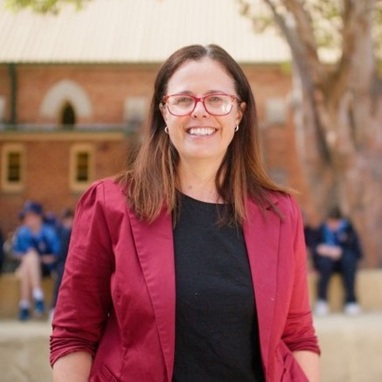 Woman with brown hair and glasses wearing a dark pink blazer, smiling for the camera