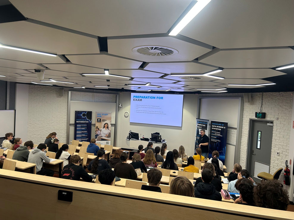 Students sit in a lecture theatre, looking towards a projecter screen that has exam preparation information