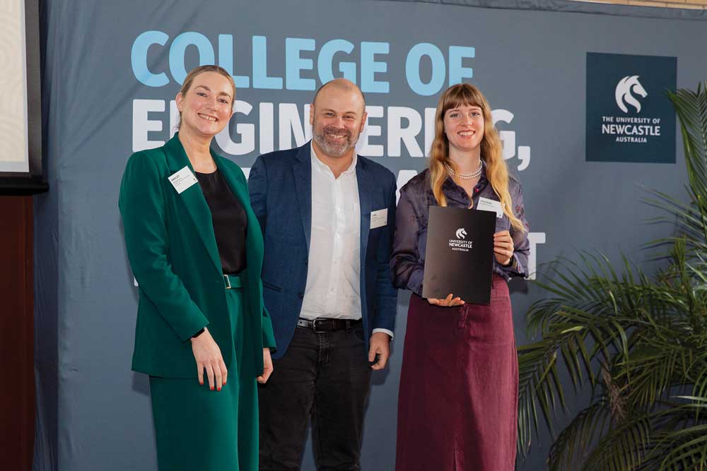 Three people pose on a stage in front of a University of Newcastle College of Engineering, Science and Environment backdrop. One person holds a University of Newcastle certificate folder while standing beside two supporters. A potted plant is visible to the side of the stage during the recognition event.