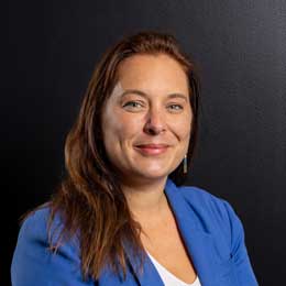 Professional head-and-shoulders portrait of Loren Collyer wearing a blue blazer over a light-coloured top, photographed against a dark background. Loren is facing the camera, with long hair worn down.