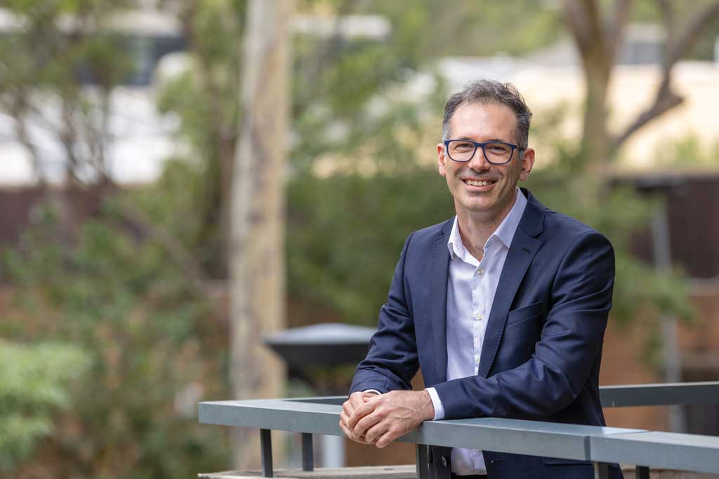 Lachlan King wearing glasses, a navy suit and a light-coloured shirt, stands leaning on a railing outdoors, with trees and landscaped greenery softly blurred in the background.