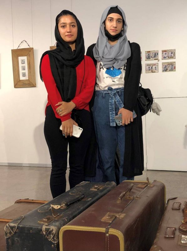 two young women in headscarves standing in front of two suitcases which are an exhibit in an art gallery