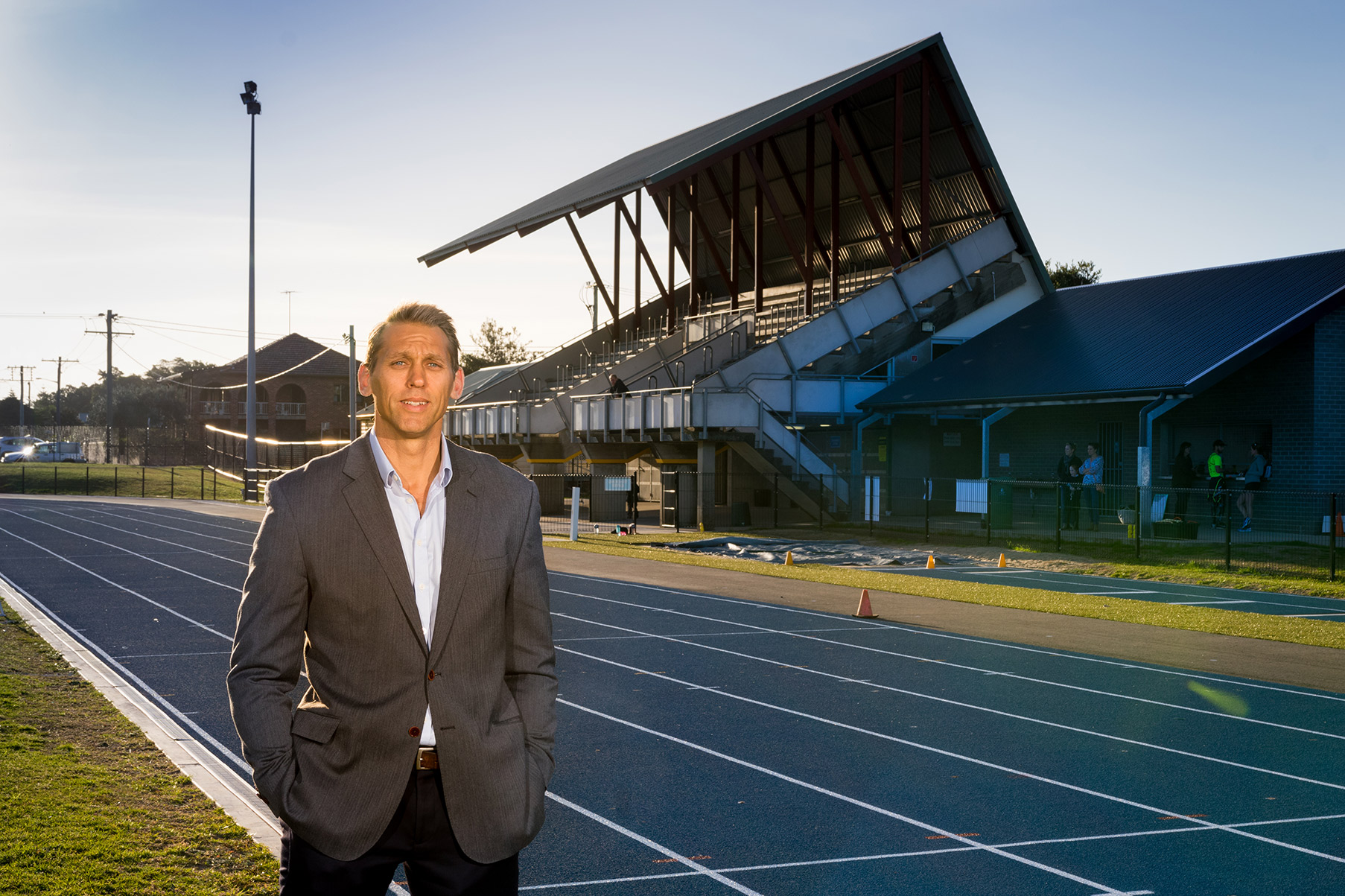 David Lubans stands on a running track with a stadium in the background looking at the camera. He is wearing a suit jacket and has his hands in his pockets