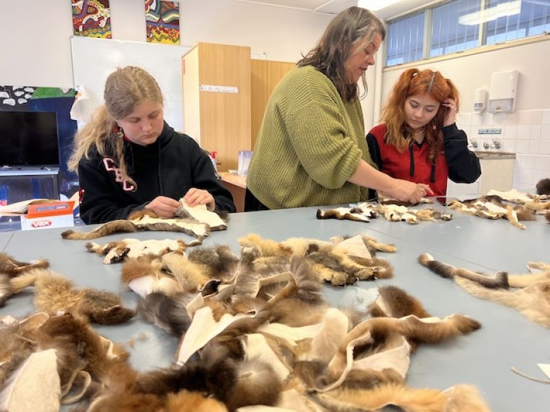 Two high school students work at a table stitching  possum skins together, pile of skin off-cuts in foreground