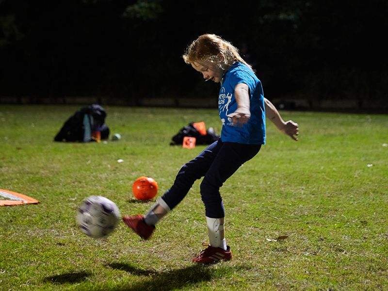 A young girl in a blue branded Daughters & Dads t-shirt mid kick of the soccer ball