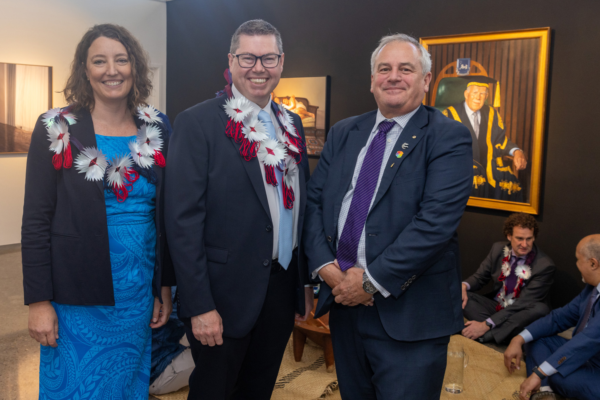 A woman in a blue dress stands next to two men wearing suits. The man in the middle has a flower lei and wears glasses. The man on the right has a purple tie. There are two men sitting on the ground behind them and a portrait of the Chancellor in the background