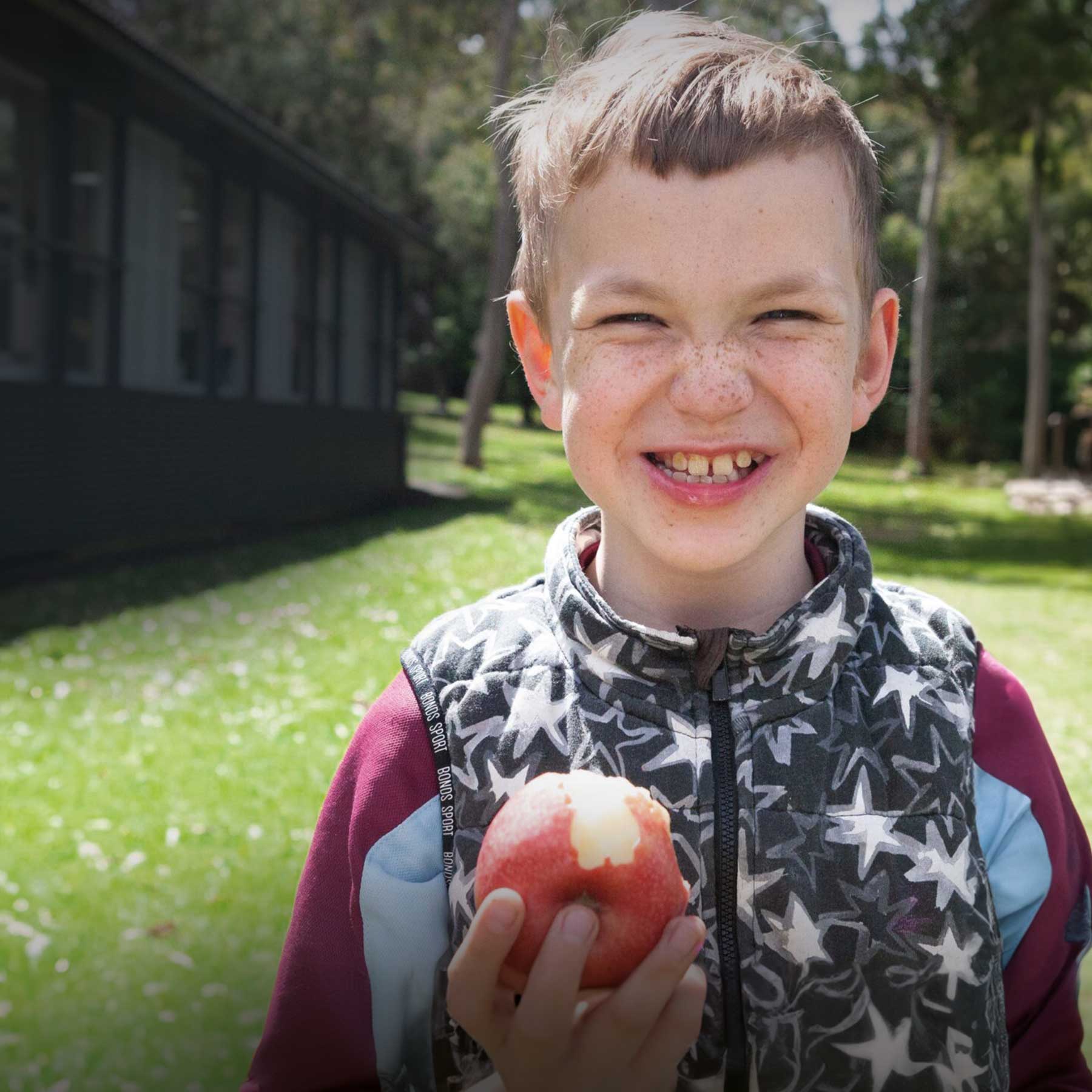 Boy standing outside, holding an apple and smiling at the camera