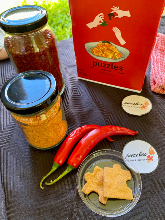 Ingredients on two jars on a table, alongsie a red box with "puzzles" written on it, two chillies, round pieces of paper with "puzzles" written on them, and two puzzle piece-shaped biscuits on a plastic lid