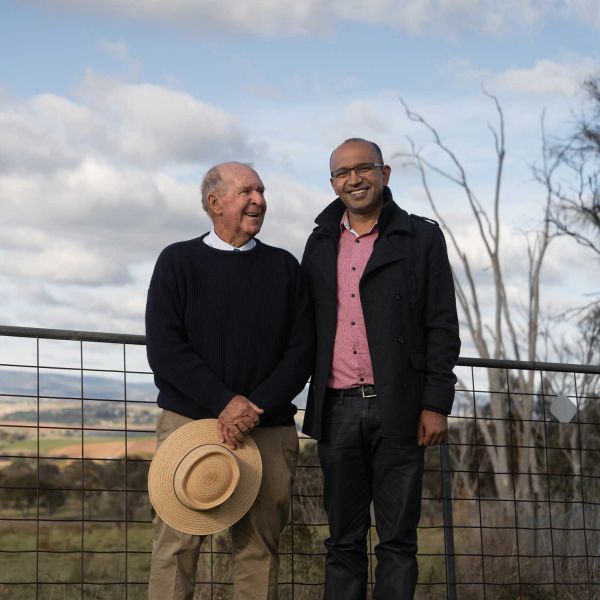Two male friends smiling for a photo in the countryside. There is a wire fence in the background