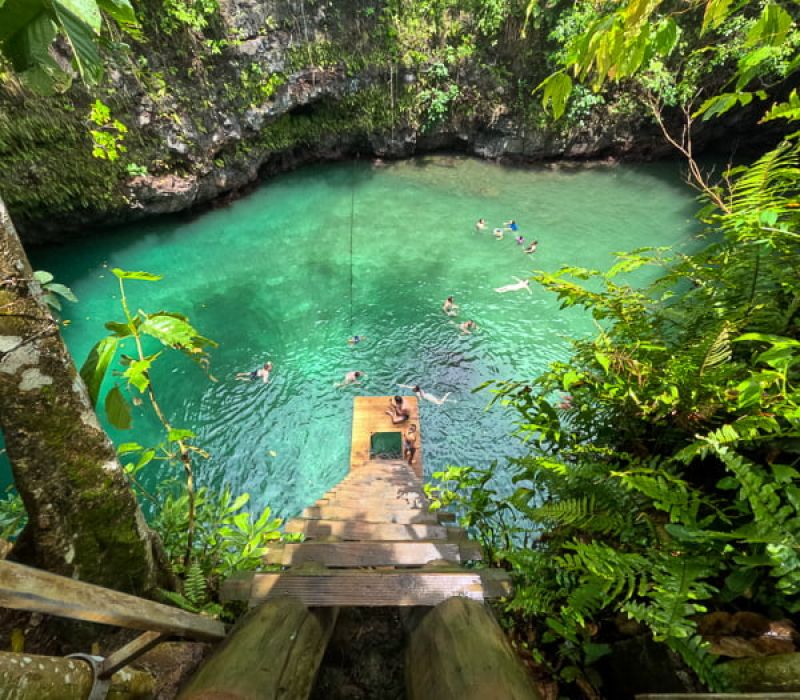 aerial view of people swimming in a large ocean trench that looks like a deep cave with clear, green water surrounded by natural rock