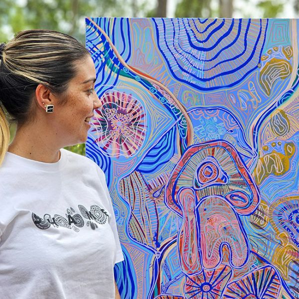 Renae stands in front of her work, titled Bulaguy Miranggul (meaning Saltbush). She is smiling, with her head on angled in profile, and looking at the artwork. She has a ponytail and white shirt on.