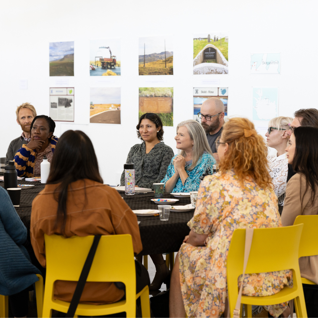 A diverse group of adults sit around a table in a bright, modern room, engaged in discussion. The table is covered with a black cloth and has drinks, snacks, and notebooks on it. Yellow chairs surround the table. Behind them, a white wall displays colorful photos and maps.