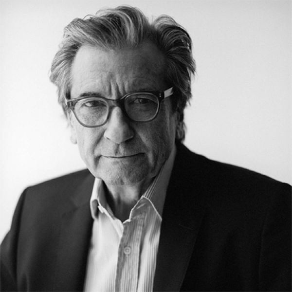 A person dressed in a dark suit jacket and collared shirt stands against a plain, light-colored background staring into the audience