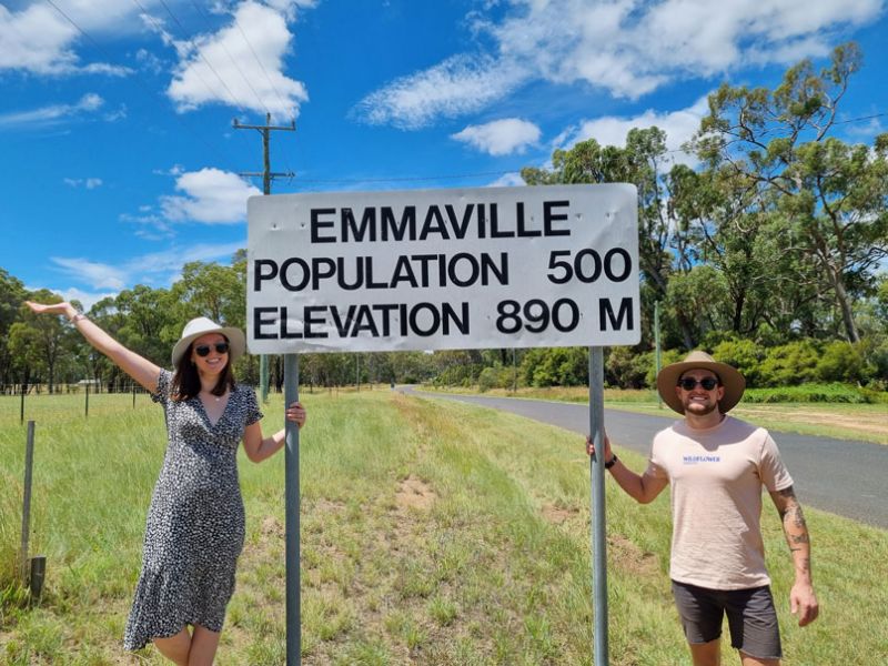 Josh stands with his partner beside a road sign that reads "Emmaville, Population 500, Elevation 890 M," with a sunny, tree-lined background.
