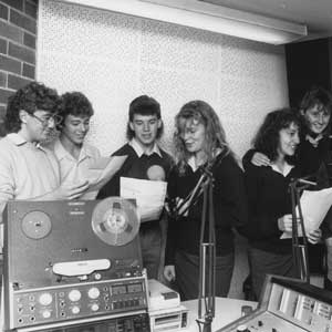 Students standing around vintage radio equipment, singing and smiling