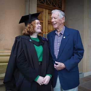 A female graduate in black cap and gown, with a green shirt underneath, stands outside a wooden door with an older man standing right next to her. They are looking at each other and smiling; he is her grandfather.