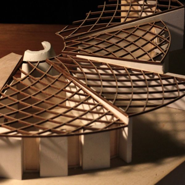 Architectural model of a building with a circular base, featuring a radial wooden lattice roof design. The structure is lit warmly, casting shadows.