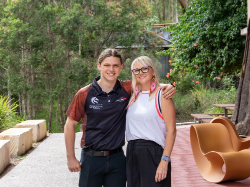 A woman and a young man stand side by side outdoors at a University campus, with trees and campus seating behind them.