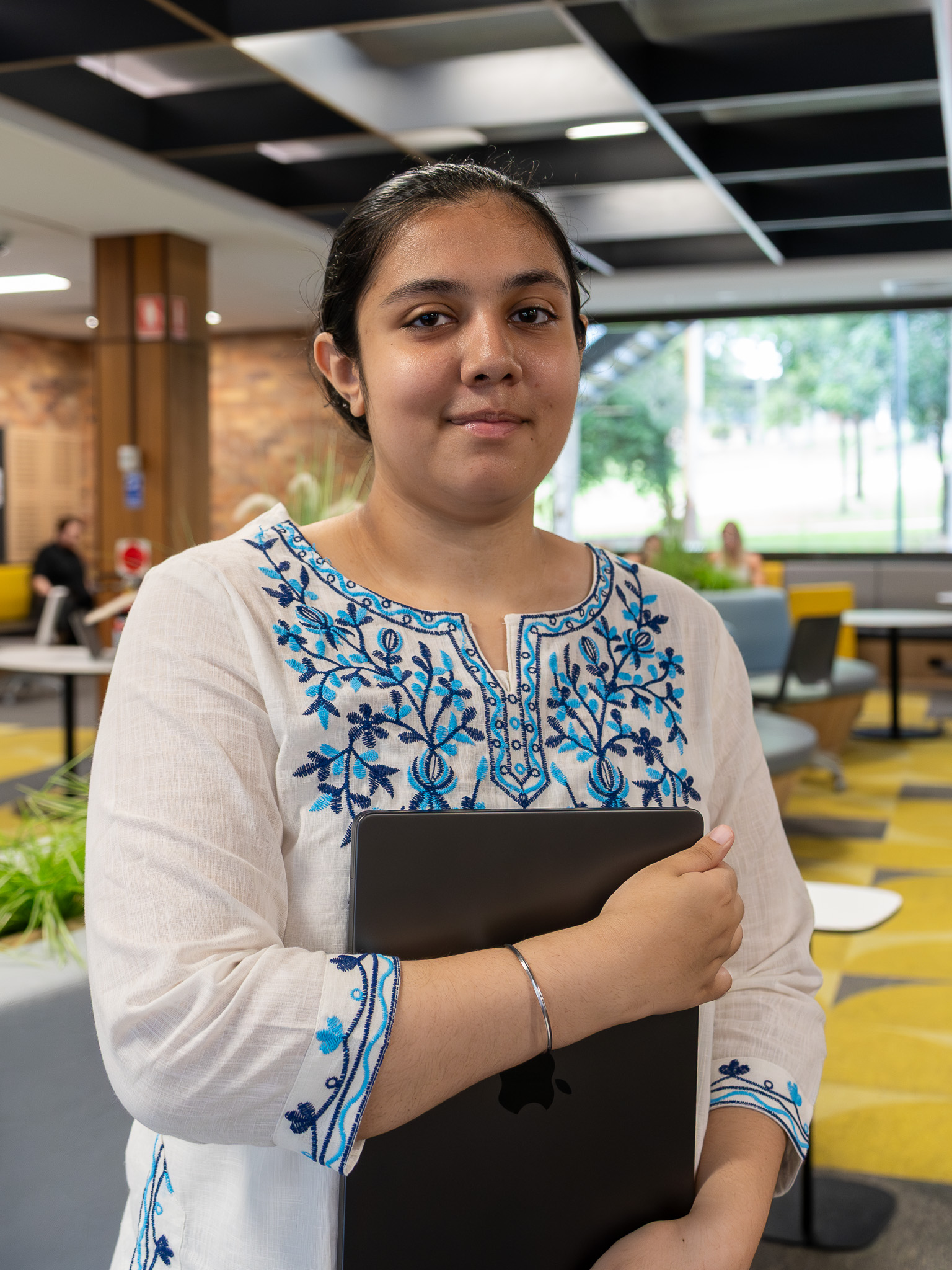 A person stands indoors in a modern university study lounge, holding a closed laptop against their chest. They are wearing a white three‑quarter‑sleeve tunic with intricate blue floral embroidery along the neckline, sleeves, and front. The background shows yellow‑and‑grey seating, large windows looking out to greenery, and several people working or talking in the space.