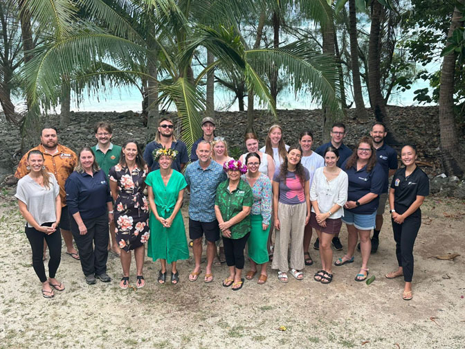Staff, students, and Cook Islands residents stand together on a beach, smiling at the camera.