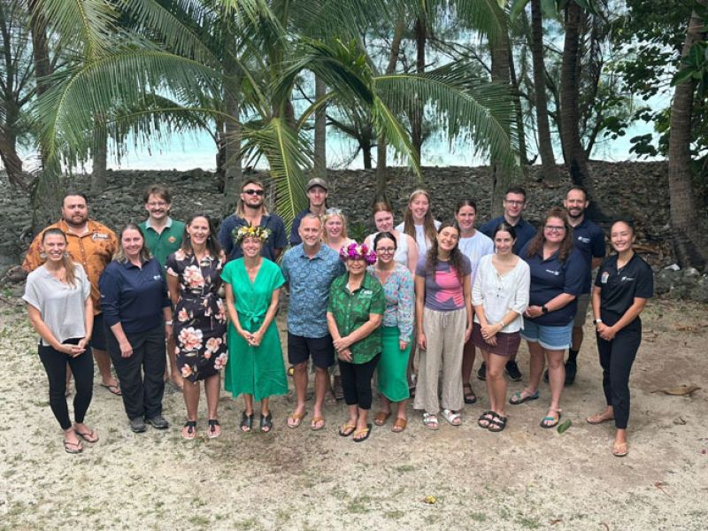Staff, students, and Cook Islands residents stand together on a beach, smiling at the camera.