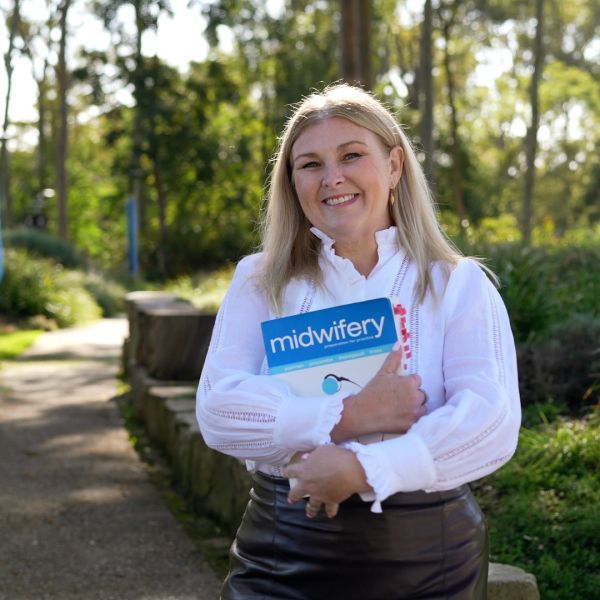 Female with blonde hair wearing a white long sleeved shirt standing outside smiling and holding a Midwifery text book.