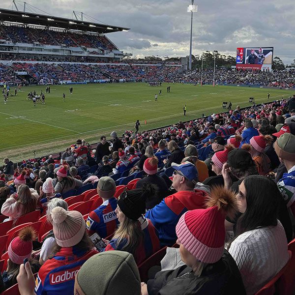 A wide crowd shot at the NRL Beanie for Brain Cancer Round in Newcastle showcasing crowds of fans, many wearing MHF Beanies, watching a game. On the big screen, two players are seen wearing beanies