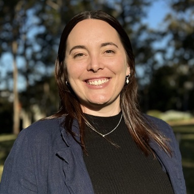Woman with dark hair, standing outside. smiling for the camera