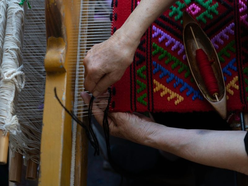 A person operates a loom, intricately weaving colorful threads into a piece of fabric.