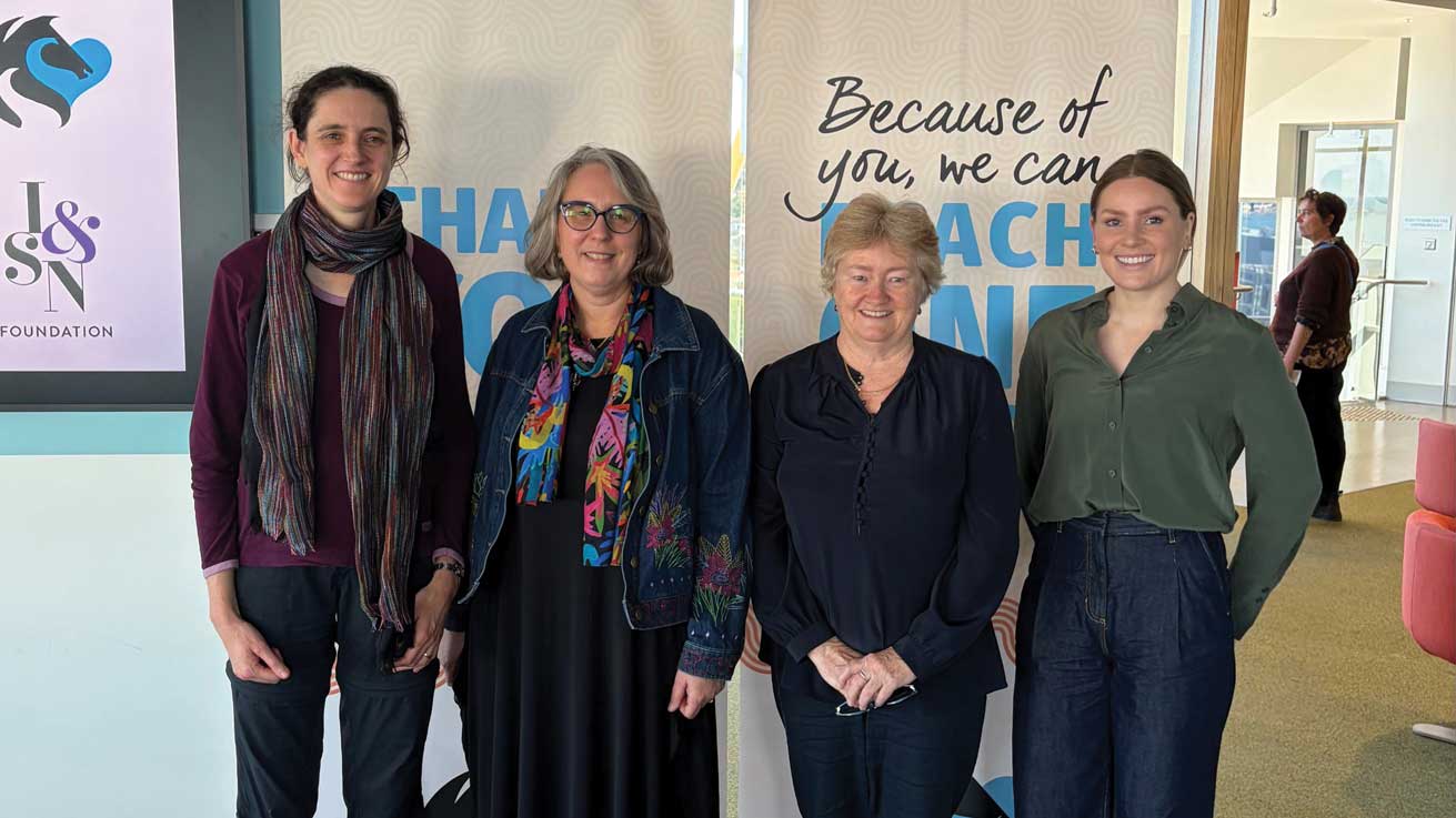 Four people stand together indoors in front of banners displaying the message “Because of you, we can reach one more”.